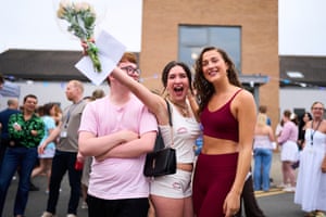 A teenage girl holds up flowers and looks delighted while stood next to two friends outside their college