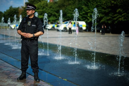 Arif Nawaz stands by a line of fountains opposite Ashton Old Baths. He wears a protective vest and peaked cap, and has his hands clasped together in front of him. He has a short beard and wears glasses.
