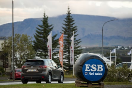A hay bale beside a road in the Reykjavik area is covered in black plastic with a sign on one side that reads 'ESB nei takk', or Europe no thanks. A dark grey SUV is being driven past and there are tall coniferous trees and misty hillsides in the background.