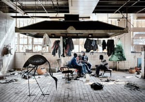 A large tiled room, with a huge machine on the ceiling (extractor fan?), from which hangs laundry. Four people talk together on mismatched chairs, surrounded by industrial detritus