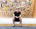 A man in a black T-shirt sitting in front of a bouldering wall.