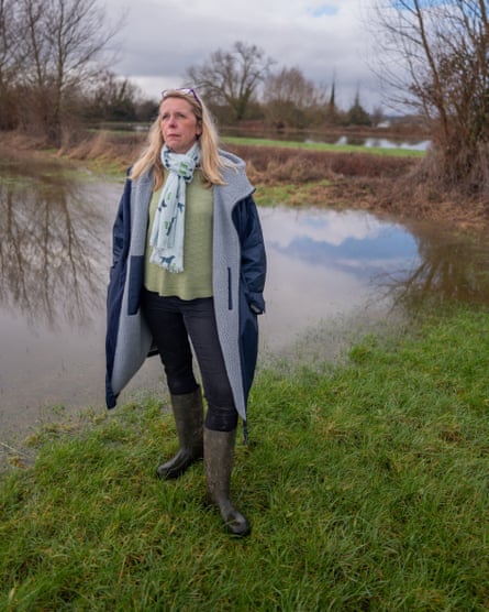 Bryony Sadler stands on grass by what looks like a curving stream but may be a large pool of flood water; trees and clouds are reflected in the brownish water; she wears wellies and a long fleece-lined coat and has long blond hair.