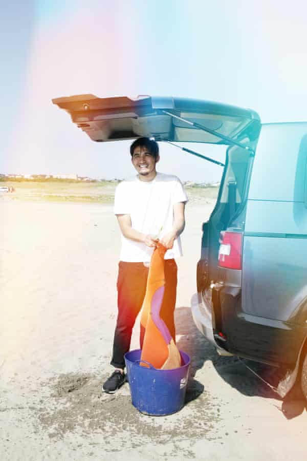 A male surfer rinsing his wetsuit out in a blue bucket while standing under the open boot of his van for sun protection