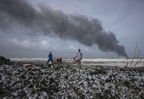 Three people walk on a beach carrying bags as a plume of smoke is seen in the distance at sea