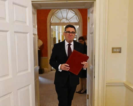 Andy Burnham walks through a doorway in a dark suit and glasses, holding a red folder