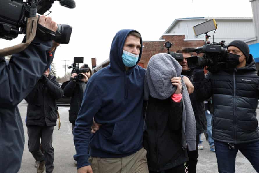A man wearing a blue hoodie and face mask walks with his arm around a woman who is shielding her face with a gray sweater from the photographers surrounding the couple.