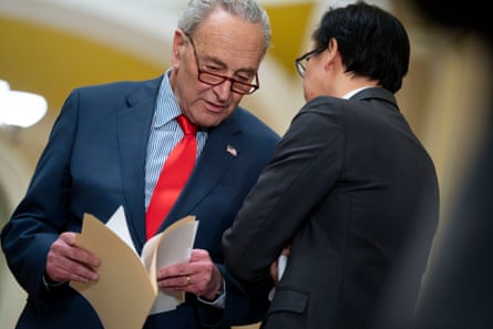 Older white man, gray hair, reading glasses, dark suit, red tie, holds manila envelope and talks to another white man in a suit.