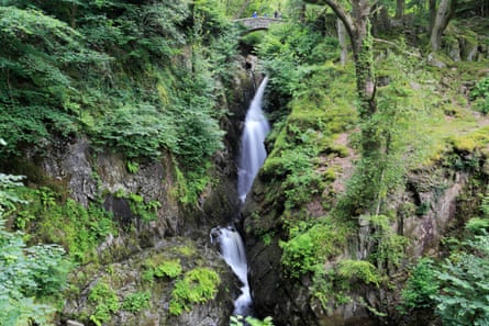 A waterfall flows down through rocky terrain, with a tiny bridge at the top showing its sheer scale