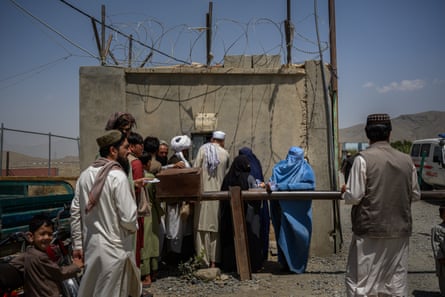 13 or so people wait outside a basic concrete hut with barbed wire on top in a desert-like landscape. The men wear shalwar kameez, some in turbans; the women wear burqas
