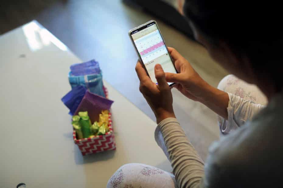 A woman sits holding her phone which is displaying a period tracking app. On the low table before her is a basket full of menstrual products.