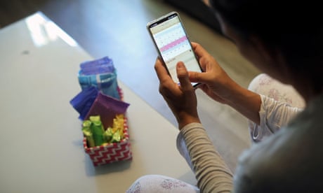 A woman sits holding her smartphone. On a low table in front of her is a small basket of feminine hygiene products.