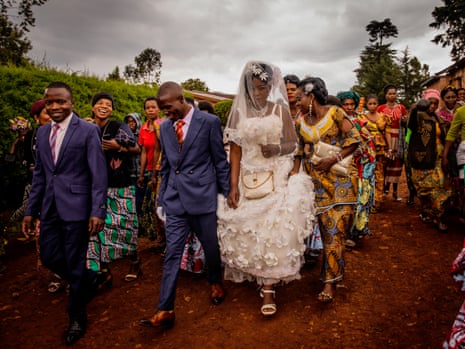 A man smiles shyly as he walks while holding the hand of a woman in a wedding dress as others crowd around