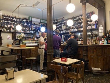 Men standing by a wooden bar area with shelves of wine behind it and small Formica tables with wooden chairs in the foreground