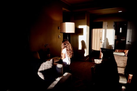 In glaringly bright sunshine coming from behind the frame, a little girl with long brown hair faces away from the camera, in a clean, neat hotel room of mostly brown and cream colors.