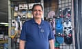 A middle-aged Latino man wearing a navy blue polo shirt with reading glasses tucked into the neck, smiles standing in front of a rack with skateboards, an electronic fly-swatter, and electronics.