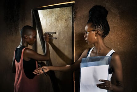 A young woman in a hut holding notes gestures as another young woman stands by a metal door about to look through a peephole 