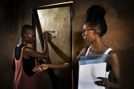 A young woman in a hut holding notes gestures as another young woman stands by a metal door about to look through a peephole