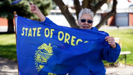 A woman wearing sunglasses and a bright blue jacket waves the Oregon state flag.