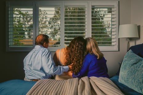 A brown-haired woman sits on the edge of a bed in between her parents, who embrace her
