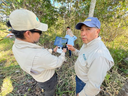 Two men in the uniform of the Rainforest Alliance, wearing caps, stand in a lightly forested area.