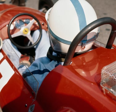A driver, shot from behind, wearing a blue and white helmet sits in a red sports car