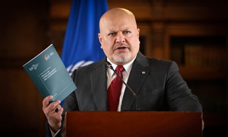 Karim Khan, speaking from behind a wooden lectern; he is holding up a policy document as he speaks and there is a blue ICC flag behind him. He is in his mid-50s and is thick-set with a bald head, dark eyes and neatly-trimmed grey beard; he wears a dark grey suit, white shirt and red tie.