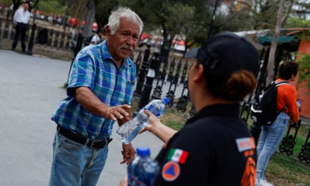 A woman in black civil protection uniform hands a bottle of water to a white-haired man in checked shirt and jeans