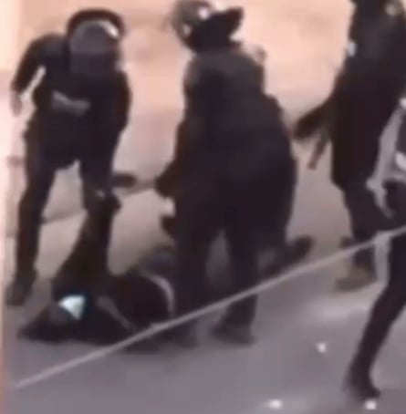 A grainy image of riot police standing over a body on the street with one pulling an arm