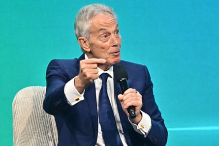 Tony Blair speaks in Vienna in June: he is gesticulating with one hand. He wears a dark blue suit and has short grey hair; he sits in front of a light turquoise-coloured screen or background.