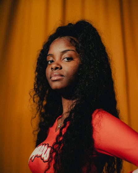 Black teenage girl, with long curly black hair down her back and wearing a long-sleeved orange T-shirt, stands in front of an orange curtain and stands sideways, looking at the camera with one hand on her hip.