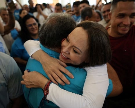 Dressed in a white long-sleeved top and surrounded by supporters, María Corina Machado smiles as she embraces a man in a blue polo shirt