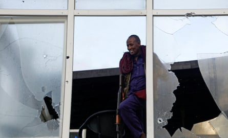Man with blue shirt and trousers looks out from window frame with smashed glass either side.