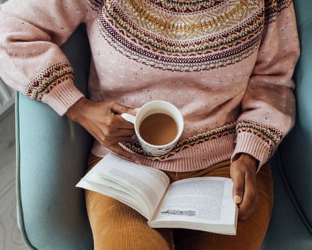 Overview view of a woman in a jumper seated in an armchair while reading a book and with the other hand holding a mug of coffee