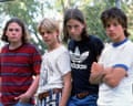 Four boys in T-shirts, two with shoulder-length hair, stand in a row in front of a chain-link fence scowling
