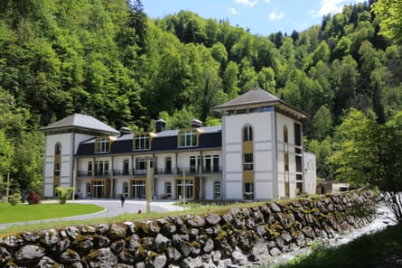 An impressive whitewashed building with forest behind and a river with stone banks running to one side