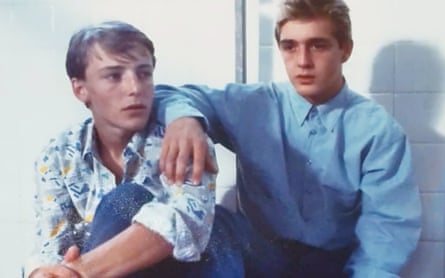 Two young men (or older teens), sitting in a locker room, one with his elbow resting on the other’s shoulder