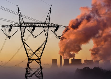 Power lines and a tower are seen in front of the Loy Yang A and B power stations in the background. There fog surrounding trees on the ground and plumes of smoke coming from the power stations