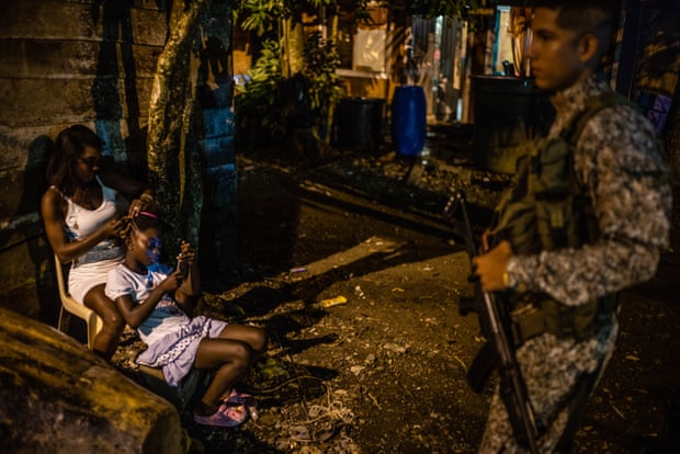 As a soldier patrols, a young woman combs the hair of a child on a street in Buenaventura