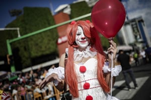 A woman wearing white face paint, with red lips and nose, holds a red balloon