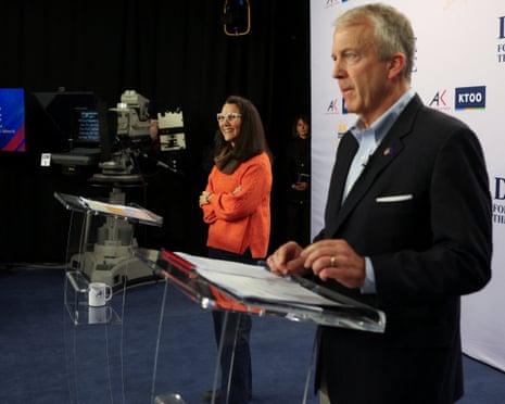 Man in suit and woman in sweater stand at lecterns near cameras during debate