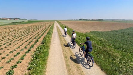 Three cyclists seen from behind take a grassy track through flat fields towards blue skies