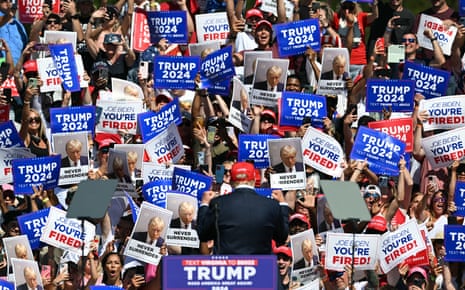 A man in a black suit and red baseball cap is seen from behind, with a sea of people cheering and holding red, white and blue Trump 2024 signs taking up most of the frame.