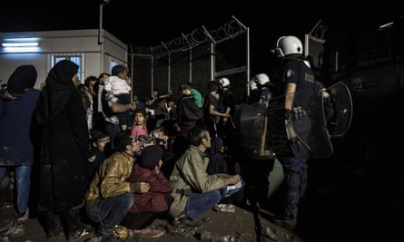 A registration camp on the Island of Lesvos at nightfall, with a group of refugees tired and frustrated sitting in the cold waiting to be processed, armed police standing over them