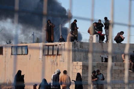People, mostly young men but also some women, stand in front of and on top of a breeze-block building as thick black smoke billows behind. They are seen through wire fencing.
