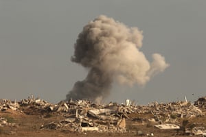 A large cloud of smoke rises in the distance behind a landscape of razed buildings