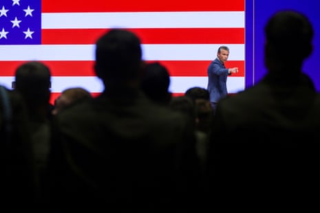 Pete Hegseth gestures on stage before a large flag screen, watched by silhouetted audience