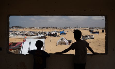 Two Palestinian children are seen silhouetted as they look through a rectangular hole in a wall at a camp for displaced people: an expanse of sand and tents are seen through the hole, including one with a sign saying Kindergarten