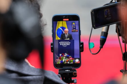 A smartphone screen shows President Lula da Silva speaking at a podium, filmed beside camera equipment