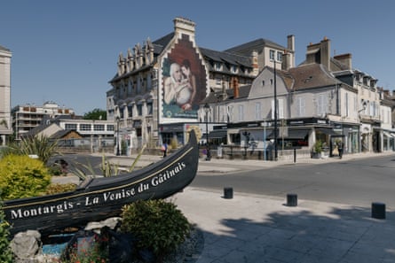 A gondola with the words ‘Montargis – La Venise du Gâtinais’ on it forms part of a small landmark on a pavement, opposite a parade of suburban shops and cafes