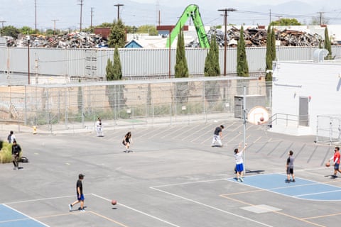 Students play basketball on an outdoor court in a schoolyard. Just beyond the fence, tall mounds of scrap metal can be seen from a recycling plant.